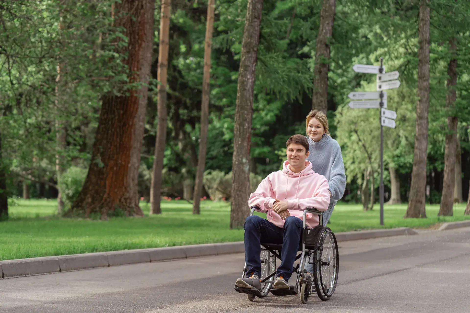 A woman assisting a young man using a wheelchair along a park path